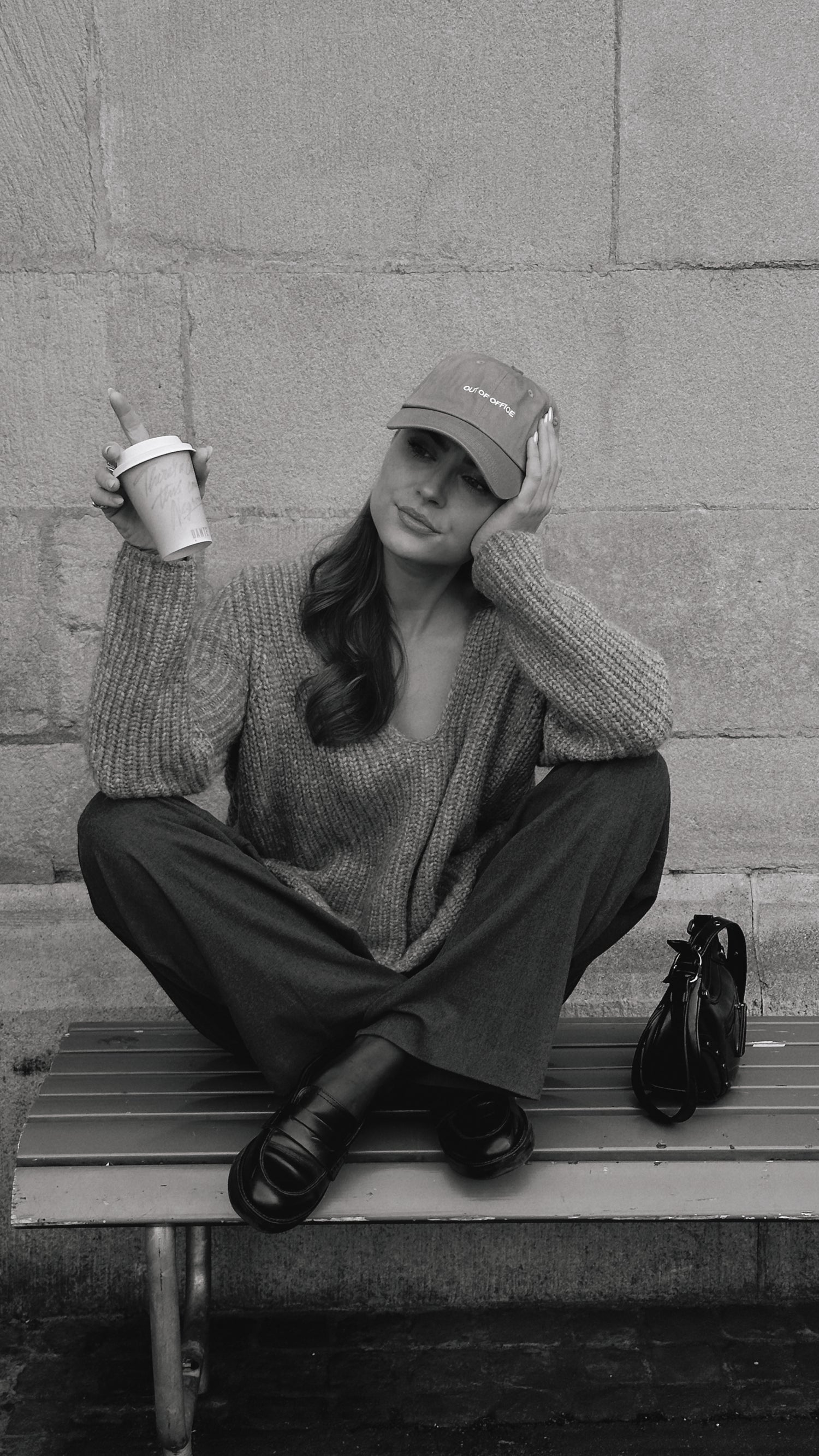 Frau sitzt auf Bank mit Kaffee, grauem Outfit und Out of Office Cap. 